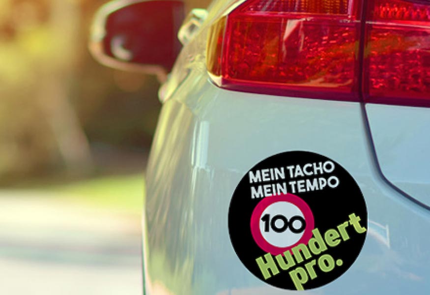 Heck eines weißen Autos mit rundem Aufkleber mit der Aufschrift "Mein Tacho, mein Tempo – Hundert pro"